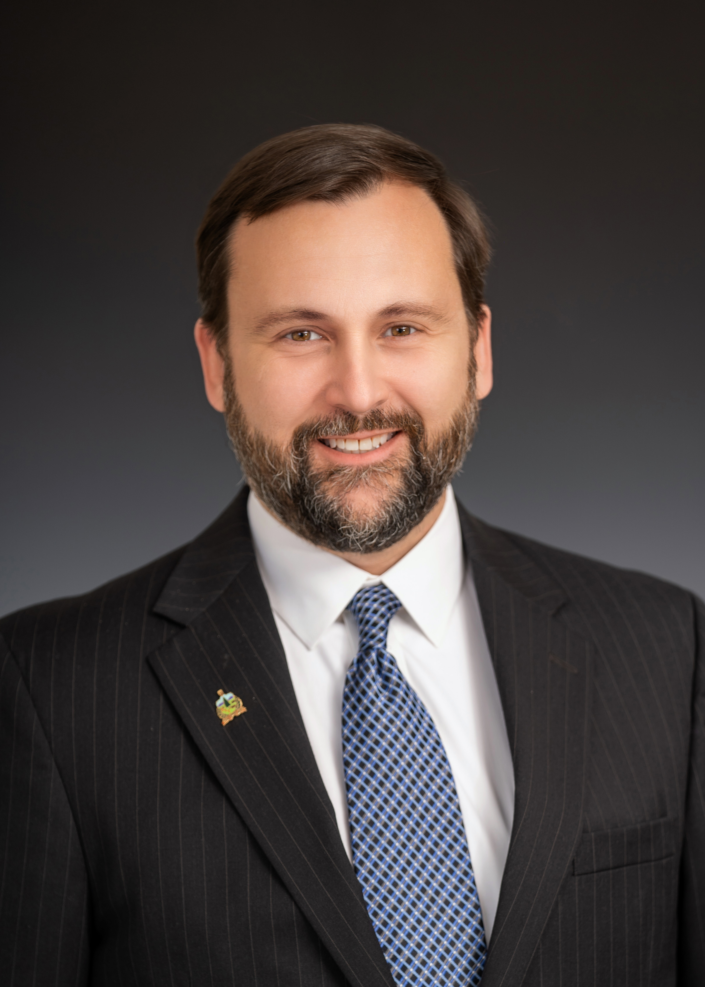 White male with dark hair and beard in black suit, white shirt and medium blue tie